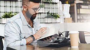 Hipster man in glasses is sitting in cafe, reading notes in notebook. On table is laptop,cup of coffee, instant camera.