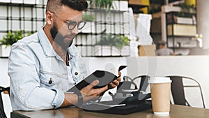 Hipster man in glasses is sitting in cafe, reading notes in notebook. On table is laptop,cup of coffee, instant camera.