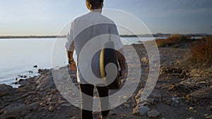 Hipster man with guitar enjoy sunset at lake coast walking on the stony beach against sun - slow-motion back view