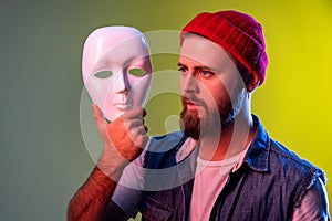 Hipster man holding white mask in hands, looking away with attentive look, hiding personality.