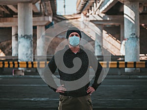 A Hipster Man In A Medical Mask For Protection Against Flu Virus Or Coronavirus Outdoor On The Background Of The Bridge. Pandemic