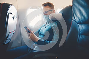 Hipster man in protection mask is watching video on mobile phone, reading online book traveling on airplane