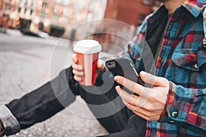 Hipster man resting with cup hot coffee on city sidewalk and texting massage on mobile phone screen photo