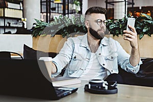 Hipster man sits in cafe, uses smartphone, works on laptop. Businessman reads an information message in phone