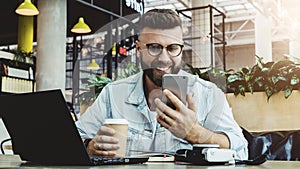 Hipster man sits in cafe,uses smartphone, works on laptop. Businessman reads information message in phone, drinks coffee