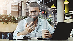 Hipster man sits in cafe,uses smartphone, works on laptop. Businessman reads information message in phone, drinks coffee