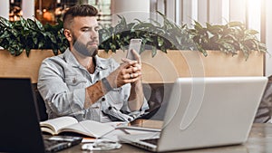 Hipster man sits in cafe, uses smartphone, works on two laptops. Businessman reads an information message in phone.