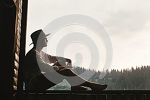 Hipster man sitting on porch of wooden house looking at mountai