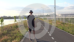 Hipster man stands with longboard, moving and playing, wearing sunglasses and hat at sunset time with skyline of city