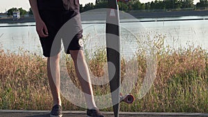Hipster man stands with longboard at waterfront in sunglasses and hat at sunset time. Summer leisure concept. Active