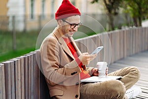 Hipster man using smartphone, chatting in social networks, sitting outdoors, drinking coffee or tea.