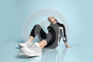 Hipster person in cap, black tracksuit, bracelet and sneakers. He is sitting on floor, posing against blue background. Close up