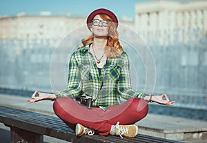 Hipster redhead woman in hat and glasses doing yoga on the bench