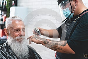 Hipster senior man getting hair cut at vintage barber shop - They both wear face protective masks for coronavirus prevention -