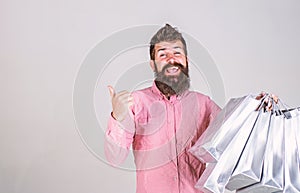 Hipster on smiling face recommends to buy. Man with beard and mustache carries bunch of shopping bags, grey background