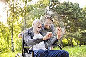 Hipster son and father in wheelchair at park taking selfie.