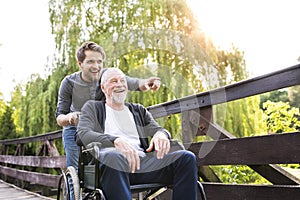 Hipster son walking with disabled father in wheelchair at park.