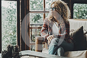 Hipster style pretty lady sitting at home on the sofa and looking outside thw windows a green forest woods. Cozy cabiness chalet