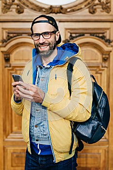 Hipster tourist in glasses, cap and yellow anorak holding backpack and smartphone having excursion in art gallery making photos be