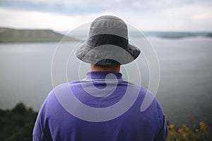 Hipster traveler in purple shirt  and bucket hat standing on top of rock mountain with amazing view on river. Young camper guy