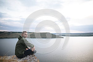 Hipster traveler sitting on top of rock mountain and enjoying amazing view on river. Stylish guy exploring and traveling.