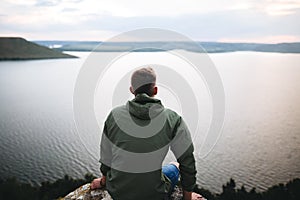 Hipster traveler sitting on top of rock mountain and enjoying amazing view on river. Stylish guy in green windbreaker exploring