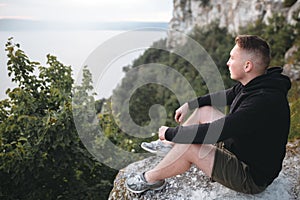 Hipster traveler sitting on top of rock mountain and enjoying amazing view on river. Stylish guy in windbreaker exploring and