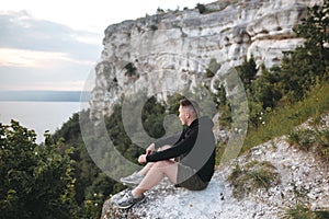 Hipster traveler sitting on top of rock mountain and enjoying amazing view on river. Stylish guy in windbreaker exploring and