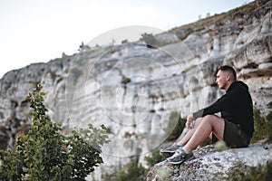 Hipster traveler sitting on top of rock mountain and enjoying amazing view on river. Stylish guy in windbreaker exploring and