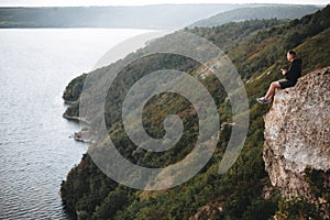 Hipster traveler sitting on top of rock mountain and enjoying amazing view on river. Young guy exploring and traveling.