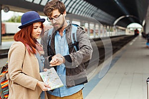 Hipster Traveller couple looking at smart watch while waiting for the train at railway station. Autumn time. Woman