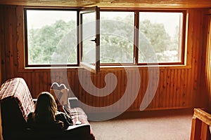 hipster woman sitting on coach and reading book in wooden cottage with view on woods, summer vacation concept, space for text