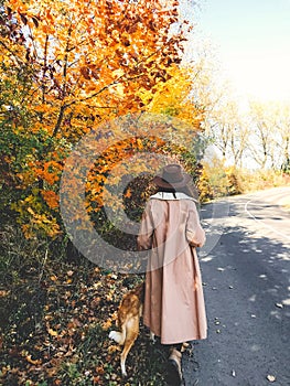 Hipster woman traveling with her golden dog, walking on autumn sunny road with fall leaves. Woman in hat and coat with dog