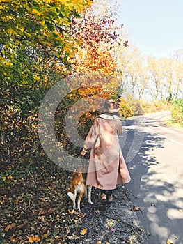 Hipster woman traveling with her golden dog, walking on autumn sunny road with fall leaves. Woman in hat and coat with dog