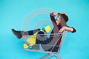 Hipster woman with yellow skateboard sitting in shopping trolley