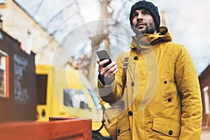 Hipster with yellow backpack, jacket, cap, coffee of thermo cup holding in the hands smartphone, freelance using gadget mobile