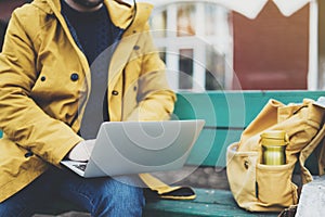 Hipster with yellow backpack, jacket, cap, thermo cup of coffee using computer open laptop in spring street outdoor, tourist man