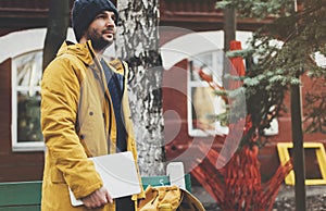Hipster with yellow backpack, jacket, cap, thermos cup of coffee holding computer laptop in spring street outdoor close up