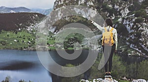 Hipster young girl with backpack enjoying sunset on peak of foggy mountain, looking on lake and poining hand. Tourist traveler