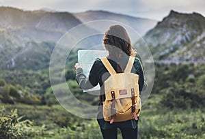 Hipster young girl with bright backpack enjoying sunset on peak of foggy mountain, looking a map and poining hand. Tourist travel