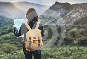 Hipster young girl with bright backpack enjoying sunset on peak of foggy mountain, looking a map and poining hand. Tourist travele