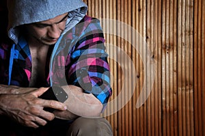 Hipster young man in the hood, sitting, holds phone and look down on wooden brown background