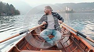 Hipster young man is paddling on the small boat in middle of magnificent lake at the background of the Alph mountains