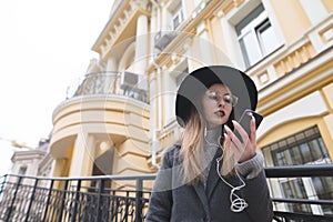 A hipsters girl with a hat and glasses looks at the phone on the background of a beautiful house.
