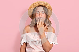 Hirizontal shot of young woman wearing summer blouse, sunglasses and straw hat, keeps hand on open mouth, being in shock, looks