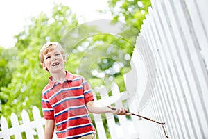 His imagination knows no bounds. A cute young boy running a stick along a white picket fence in his backyard.