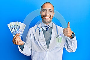 Hispanic adult doctor man wearing medical uniform holding 100 south african rand smiling happy and positive, thumb up doing