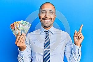 Hispanic adult man holding australian dollars smiling happy pointing with hand and finger to the side