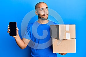 Hispanic adult man holding delivery packages and showing smartphone screen smiling looking to the side and staring away thinking