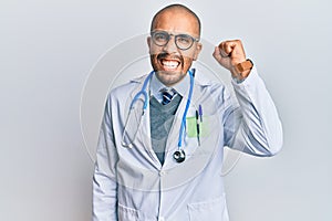 Hispanic adult man wearing doctor uniform and stethoscope angry and mad raising fist frustrated and furious while shouting with